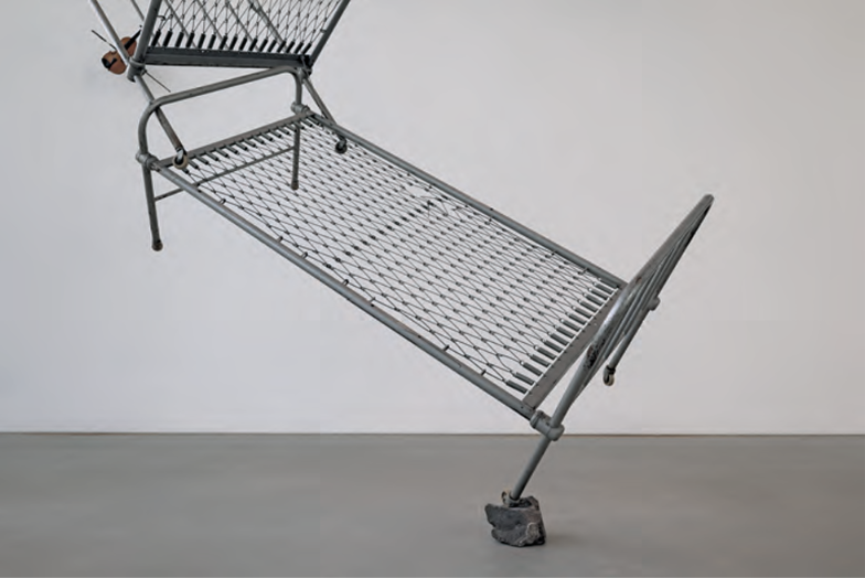 Schräges Metallbettgestell ohne Matratze, ein Bein auf einem Stein abgestützt vor weißer Wand.